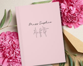 May include: A light pink notebook featuring the text Miss Sophie in black script and a black line art illustration of three ballet dancers in various poses centered on the front cover of the rectangular book.