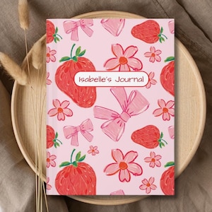May include: Pink journal cover with a repeating pattern of strawberries, bows, and flowers. The text "Isabelle's Journal" is printed in red on the cover.