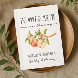 May include: A white book with the text "THE APPLE OF OUR EYE is on the way" and "WISHES FOR THE FUTURE PARENTS Sally & Harry". The cover features a watercolor illustration of apples and blossoms.