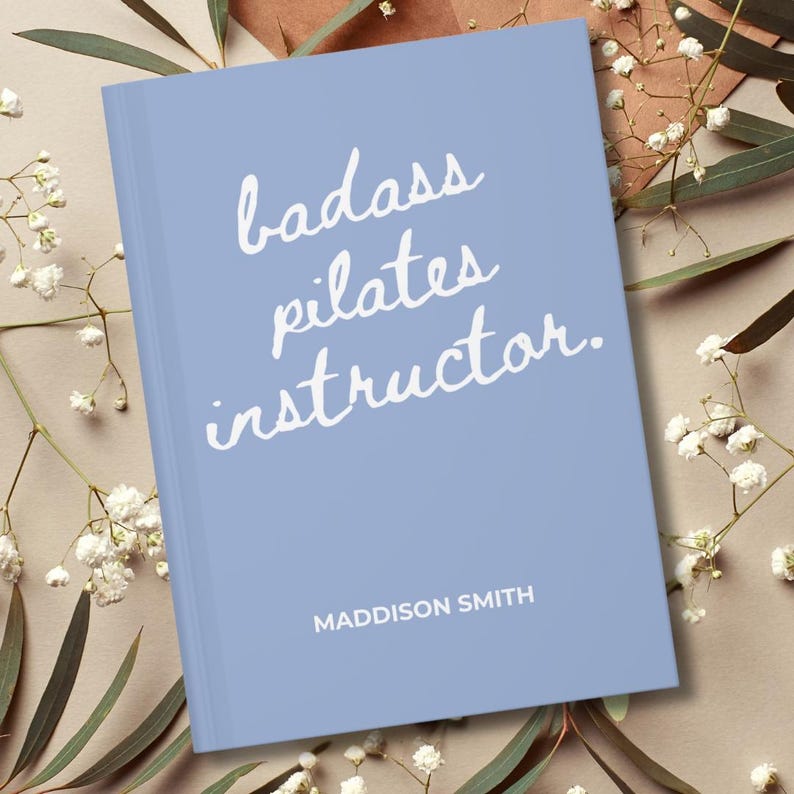 May include: A light blue book with the words "badass pilates instructor" in white script. The author's name, Maddison Smith, is printed in a smaller font at the bottom. The book is surrounded by white flowers and greenery.