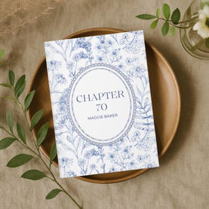 May include: A hardcover book with a blue floral design and the text "CHAPTER 70 MAGGIE BAKER" in a decorative oval. The book is on a wooden plate, surrounded by greenery, creating a natural, elegant aesthetic.