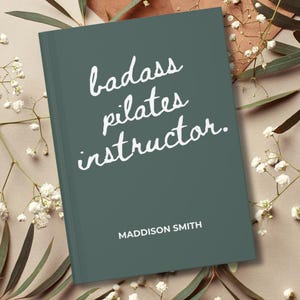 May include: A teal-colored book with the words "badass pilates instructor" in white script. The name "Maddison Smith" is printed below the title. The book is surrounded by white flowers and greenery.