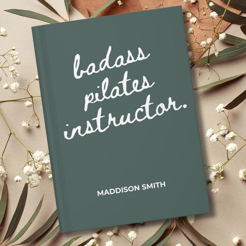 May include: A teal-colored book with the words "badass pilates instructor" in white script. The name "Maddison Smith" is printed below the title. The book is surrounded by white flowers and greenery.
