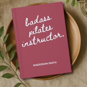 May include: A maroon-colored book with the text "badass pilates instructor" in white script. The author's name, Maddison Smith, is printed at the bottom. The book is on a wooden plate with greenery.