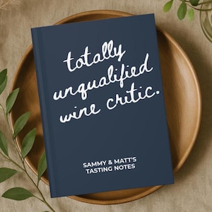 May include: A navy blue notebook with the text "totally unqualified wine critic" in white script. Below the text is "SAMMY & MATT'S TASTING NOTES" in a smaller font. The notebook is on a wooden plate.
