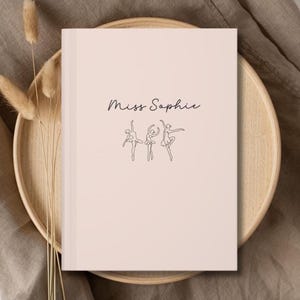 May include: A light pink notebook with the text "Miss Sophie" in cursive and three line-drawn ballerinas. The notebook is on a wooden tray with dried plants, set against a neutral background.