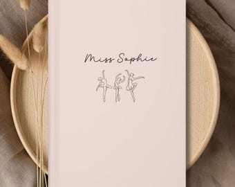 May include: A light pink notebook with the text "Miss Sophie" in cursive and three line-drawn ballerinas. The notebook is on a wooden tray with dried plants, set against a neutral background.