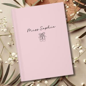 May include: A light pink journal with the text "Miss Sophie" in cursive script and a line drawing of ballet shoes. The journal is closed and has a smooth cover. The background includes white flowers and greenery.