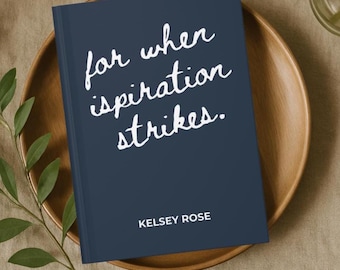 May include: A navy blue journal with the text "for when inspiration strikes" in white script. The journal is on a wooden plate, with the name "KELSEY ROSE" at the bottom. Greenery is in the background.