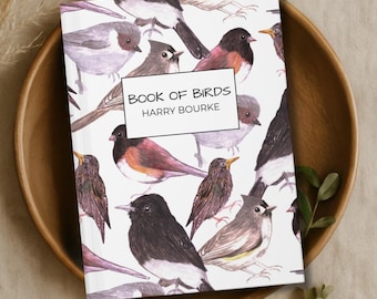Puede incluir: Un libro titulado "BOOK OF BIRDS" de Harry Bourke. La portada presenta ilustraciones en acuarela de varias aves en tonos marrones, grises y negros sobre un fondo blanco. El libro está en un cuenco de madera.