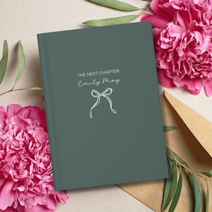 May include: A dark teal journal with the text "THE NEXT CHAPTER" and "Emily May" in white script, along with a bow illustration. The journal is surrounded by pink peonies and a brown envelope.
