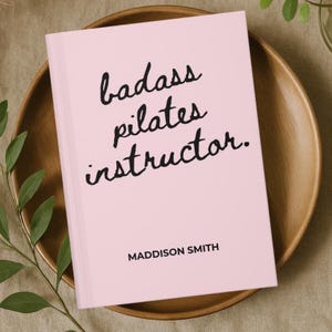 May include: A light pink book with the words "badass pilates instructor" in black script. The author's name, Maddison Smith, is printed in a simple black font at the bottom. The book rests on a wooden plate.