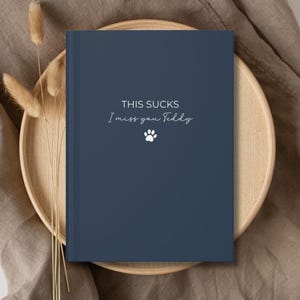 May include: A navy blue journal with the text "THIS SUCKS I miss you Teddy" and a paw print. The journal is on a wooden plate with dried flowers, set against a neutral fabric background.