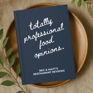 May include: A navy blue book with the text "totally professional food opinions." in white script. Below the title is "BEC & MATT'S RESTAURANT REVIEWS" in white capital letters. The book is on a wooden plate.