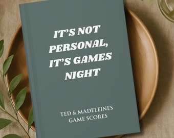 Puede incluir: Un cuaderno de color verde azulado con el texto "IT'S NOT PERSONAL, IT'S GAMES NIGHT" en blanco. Debajo, se lee "TED & MADELEINES GAME SCORES". El cuaderno está sobre un plato de madera con vegetación.