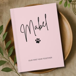 May include: A pale pink journal featuring the name "Mabel" in black script, a paw print, and the text "OUR FIRST YEAR TOGETHER." The journal rests on a wooden plate, surrounded by foliage.