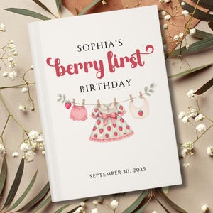 Puede incluir: Una portada de libro blanca con el texto "SOPHIA'S berry first BIRTHDAY" en escritura rosa. Debajo, una cuerda de ropa muestra ropa de bebé: una braga, un vestido con detalles de fresas y un babero. La fecha "SEPTEMBER 30, 2025" está en la parte inferior.