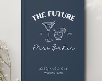 Op de afbeelding: Een marineblauwe trouwplanner met witte tekst en illustraties. De cover leest "THE FUTURE Mrs Baker" met een martiniglas en een cocktailglas. Hieronder staat "Lilly and John's WEDDING PLANS".