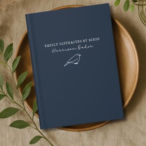 Könnte beinhalten: Ein marineblaues Notizbuch mit dem Text "EASILY DISTRACTED BY BIRDS" und "Harrison Baker" in weißer Schrift. Eine weiße Strichzeichnung eines Vogels befindet sich unter dem Text. Das Notizbuch liegt auf einem Holztablett.