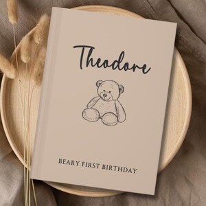 以下が含まれることがあります： ベージュ色の本。黒い筆記体で「Theodore」と書かれ、テディベアのスケッチが描かれています。ベアの下には「BEARY FIRST BIRTHDAY」の文字。本は木製のトレイの上に置かれています。