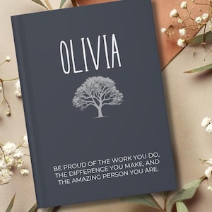 May include: A dark blue journal with the name "Olivia" printed in white. The journal features a white tree illustration and the text "Be proud of the work you do, the difference you make, and the amazing person you are."