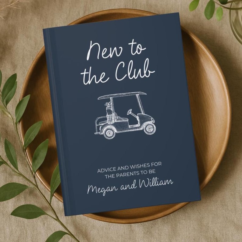 Op de afbeelding: Een marineblauw gastenboek met witte tekst met de tekst "New to the Club" en een schets van een golfkar. De tekst "Advice and Wishes for the Parents to Be" en de namen "Megan and William" zijn ook zichtbaar.