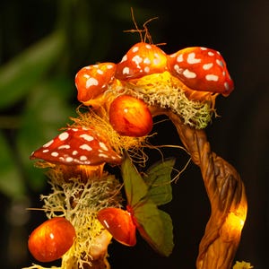 May include: A decorative mushroom lamp with a whimsical design. The lamp features a cluster of red and white mushrooms with white spots, a brown twig base, and a green butterfly. The mushrooms are illuminated with warm white LED lights.