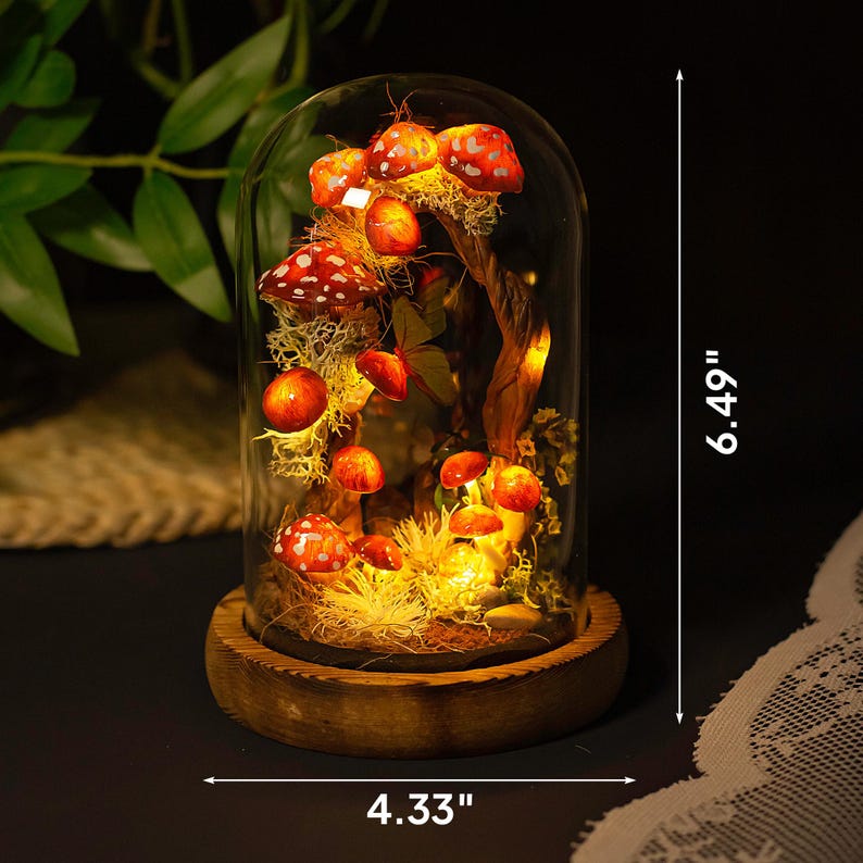 May include: A glass dome with a wooden base containing a miniature forest scene with red and white mushrooms, moss, and a small tree branch. The dome is lit from within with warm white LED lights. The dome is 6.49 inches tall and 4.33 inches wide.