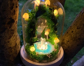 May include: A glass dome terrarium with a miniature forest scene. The scene includes a small pond with blue water, white crystals, and moss-covered ground. There are several small, white mushrooms with glowing yellow lights inside. The terrarium is on a wooden base.