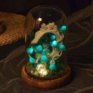 Puede incluir: Una cúpula de vidrio con una base de madera contiene una escena de bosque en miniatura con un árbol blanco, setas azules y una pequeña figura. Las setas están iluminadas con pequeñas luces LED.