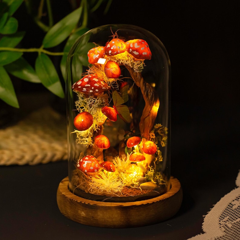 May include: A glass dome with a wooden base containing a miniature forest scene with red and white mushrooms, moss, and a twig. The scene is illuminated with warm white fairy lights.