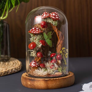 May include: A glass dome with a wooden base containing a miniature forest scene with red and white mushrooms, moss, and a twisted tree branch.