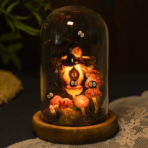 May include: A glass dome with a wooden base containing a whimsical Halloween scene.  Inside the dome are glowing mushrooms with googly eyes, a witch's hat, and a black bat.