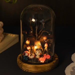 Puede incluir: Una cúpula de vidrio con una base de madera contiene una escena espeluznante de Halloween. Un árbol nudoso con ojos brillantes se encuentra en el centro, rodeado de hongos y globos oculares brillantes.