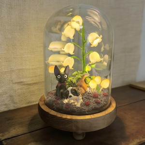 May include: A decorative glass dome with a wooden base houses a miniature scene. Inside, a glowing lily of the valley plant and mushroom lights illuminate a black cat, a dog, and a brown cat figurine. Red rose accents complete the whimsical display.