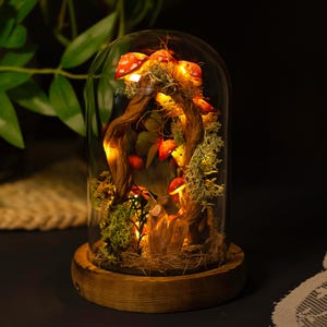 May include: A glass dome with a wooden base containing a miniature forest scene with red and white mushrooms, moss, and a twisted branch with fairy lights.