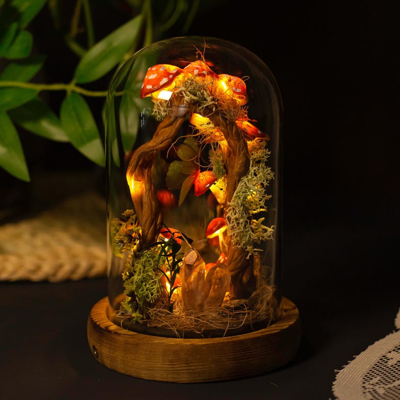 May include: A glass dome with a wooden base containing a miniature forest scene with red and white mushrooms, moss, and a twisted branch with fairy lights.