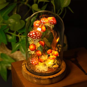 May include: A glass dome with a wooden base containing a miniature forest scene with red and white mushrooms, moss, and a small tree branch. The dome is illuminated from within, creating a warm glow.