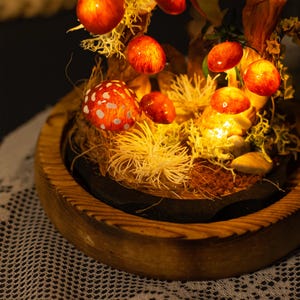 May include: A wooden base with a black circular insert holds a miniature forest scene with glowing red and white mushrooms. The mushrooms are arranged on a bed of moss and dried grasses.