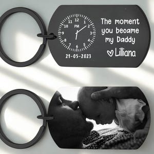 May include: Black metal keychain with two rectangular pendants. One pendant features a clock design with the text "The moment you became my Daddy Lilliana" and the date "21-05-2023". The other pendant displays a black and white photo.