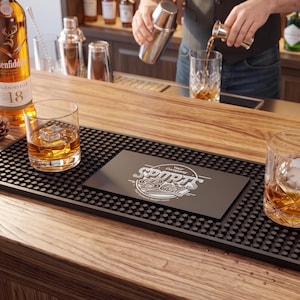 May include: A rectangular black rubber bar mat with a textured nub surface and a central smooth panel featuring the white printed text "Your Company SPORTS Bar". The mat is positioned on a wooden counter between two glass tumblers containing amber liquid.