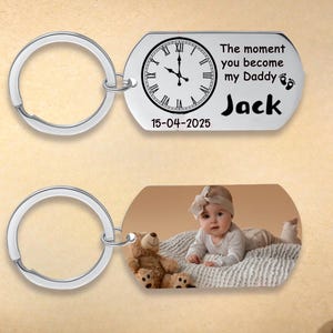 May include: Two silver keychains. One keychain has a tag with a clock face, the text "The moment you become my Daddy" and the name "Jack". The other keychain has a photo of a baby and a teddy bear.