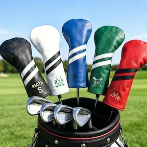 May include: A golf bag with a set of clubs and five personalised leather head covers. The head covers are black, white, blue, green, and red, each with a unique initial or monogram and stripe design. The golf clubs are silver.