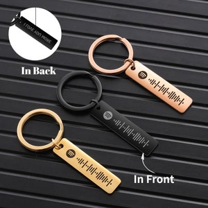 May include: Two keychains with a black and gold metal finish. Each keychain has a rectangular tag with a Spotify code and the text "I love you more" on the back. The keychains are on a black and white striped surface.