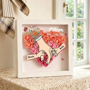 May include: A white framed artwork featuring a heart-shaped floral arrangement in pink and orange hues. The artwork includes illustrations of hands holding each other, with the names "Annie" and "Bella". The text "Mom WE LOVE YOU" is also included.