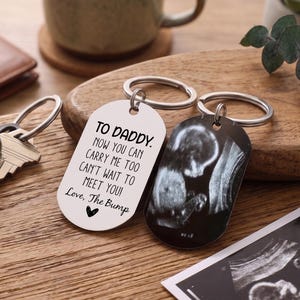 Könnte beinhalten: Zwei silberfarbene Schlüsselanhänger in Form von Erkennungsmarken. Eine Marke trägt den schwarzen Text: TO DADDY. NOW YOU CAN CARRY ME TOO CAN'T WAIT TO MEET YOU! Love, The Bump mit einem Herzsymbol. Die andere zeigt ein schwarz-weißes Ultraschallbild.