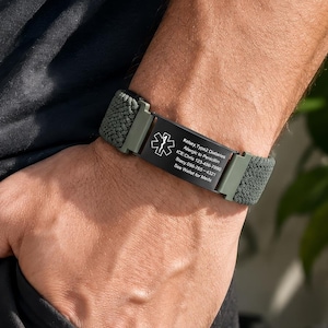 May include: An olive green medical alert bracelet featuring a woven fabric band and a black rectangular metal plate. The plate displays a white Star of Life symbol and engraved text detailing medical information and emergency contact numbers.