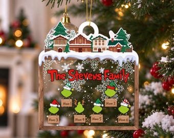 May include: A square Christmas ornament featuring a snowy village scene at the top and a clear center with the red text The Stevens Family. Below are six green cartoon characters wearing holiday hats, each holding a wooden name sign.