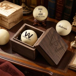 May include: A selection of personalized billiard balls and wooden boxes. One ball is inside a dark wooden box, another has the names "Paul & Josie" and the date "06.04.2026". The boxes and balls have custom text.