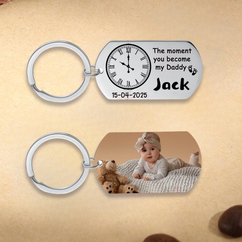 May include: Two silver keychains. One keychain has a tag with a clock face, the text "The moment you become my Daddy" and the name "Jack". The other keychain has a photo of a baby and a teddy bear.